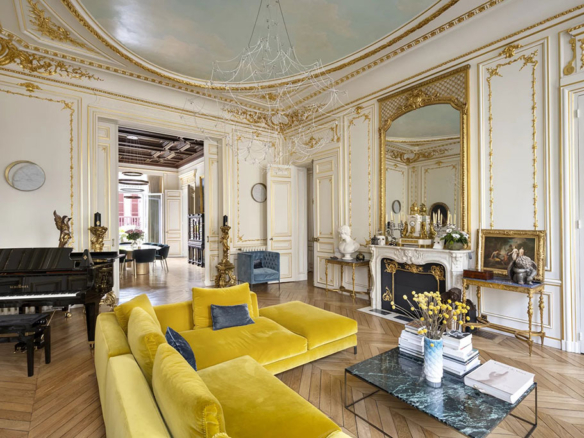 Image of an elegant living room in a luxury Paris apartment with ornate gold-trimmed walls, a grand piano, a yellow velvet sofa, and a marble fireplace with a large mirror.