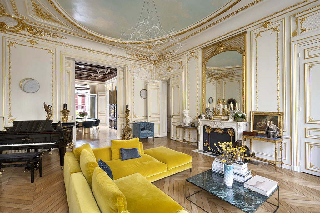 Image of an elegant living room in a luxury Paris apartment with ornate gold-trimmed walls, a grand piano, a yellow velvet sofa, and a marble fireplace with a large mirror.