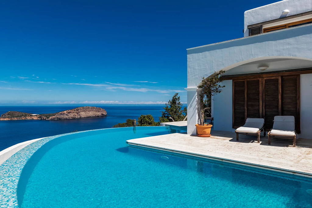 Image of a luxurious seaside villa in Ibiza with an infinity pool overlooking a calm blue ocean and a distant rocky island under clear skies.
