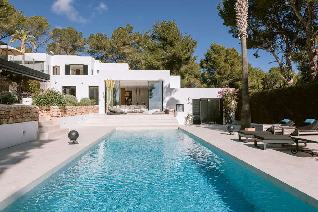 Image of a modern white villa in Ibiza with a long rectangular pool, surrounded by trees, sun loungers, and a peaceful garden setting.