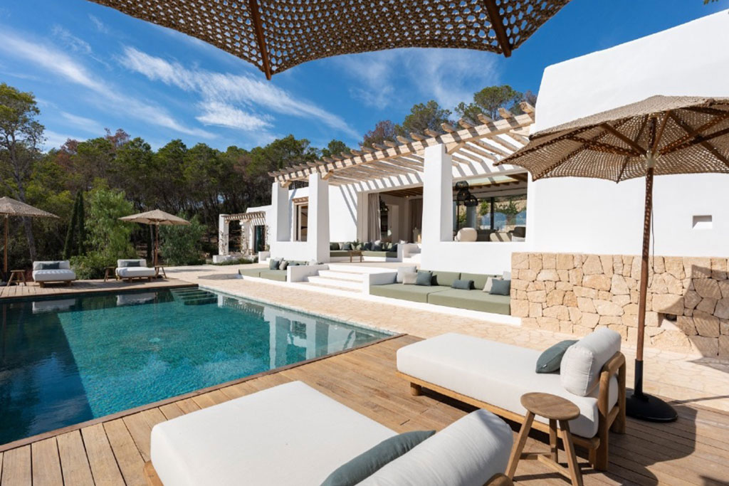 Image of a stylish Mediterranean villa with white walls, stone accents, a wooden deck, and a serene pool surrounded by cushioned lounge seating.