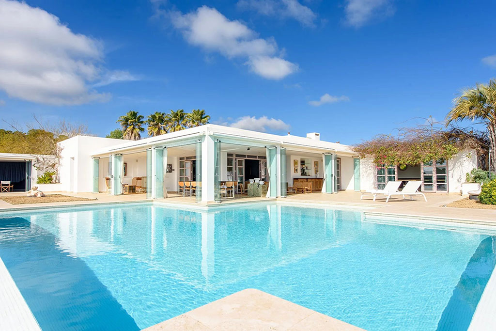 Image of a bright modern villa in Roca Llisa, Ibiza with glass doors opening onto a large turquoise pool, surrounded by palm trees and clear blue skies.