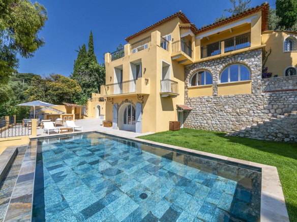 Image of a luxury french villa with stone accents, sunlit terraces, and a clear rectangular pool surrounded by lush greenery under a bright sky.