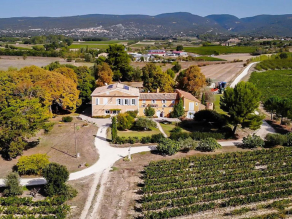 Image of an aerial view of a countryside estate surrounded by vineyards, trees, and winding paths, with rolling hills and farmland stretching into the distance.