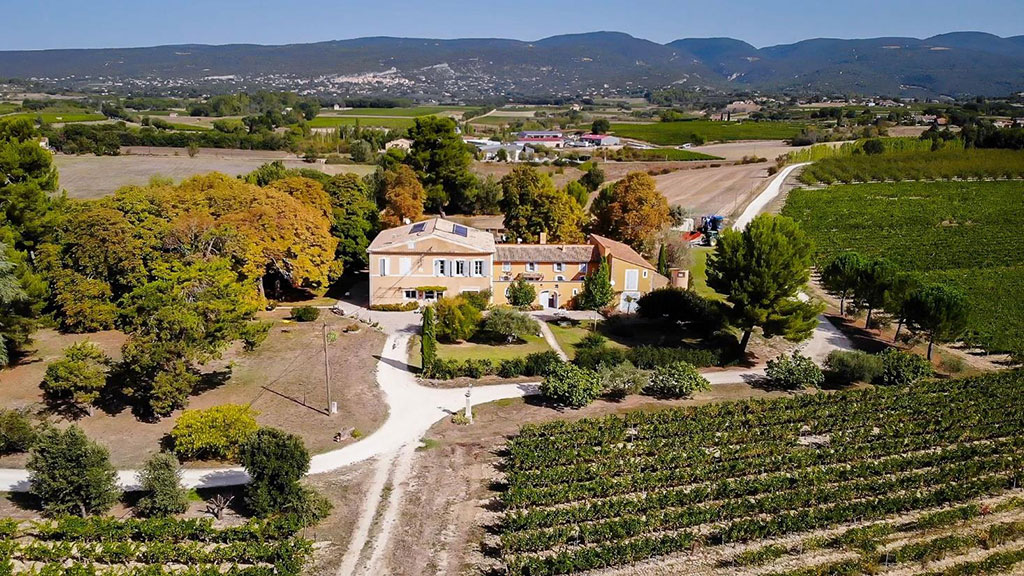 Image of an aerial view of a countryside estate surrounded by vineyards, trees, and winding paths, with rolling hills and farmland stretching into the distance.